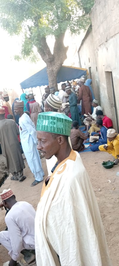 This image depicts an outdoor gathering of approximately twenty-five individuals, predominantly adult males, in Kabuga, Nigeria, under daylight conditions. The ground is unpaved and appears dusty. A large tree with a thick trunk and green foliage dominates the upper left quadrant, with a blue rectangular canopy positioned beneath it, providing shade. A weathered, light-colored concrete building wall is visible along the right side of the frame.

In the immediate foreground, a man is shown in profile, facing right, wearing a cream-colored robe with tan embroidery on the shoulders and a green skullcap adorned with white geometric patterns. He has a short beard and his gaze is directed downwards. To his left and slightly behind, a man in a light blue robe and sandals stands near an unpaved trench or depression in the ground.

The mid-ground and background feature multiple individuals, primarily men dressed in traditional robes and various caps. Several are seated on the ground or on blue mats, actively engaged in reading from open books or pamphlets. One man in yellow attire is seated on the far right, near a green bowl placed on the ground. A woman wearing a light blue head covering is also discernible within the gathered crowd. The subjects are clustered, indicating a communal assembly.