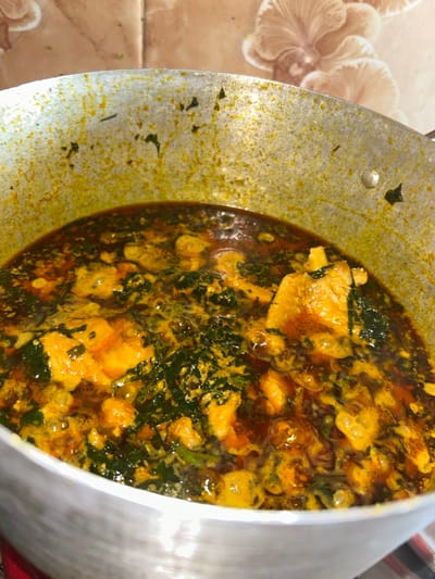The scene depicts a large, metallic cooking pot containing a prepared, simmering dish, located in Egbe, Nigeria. The pot holds a robust stew or soup, characterized by a vibrant reddish-orange oil base with visible floating oil droplets. Within the liquid, distinct elements of dark green leafy vegetables and irregularly shaped, light orange-yellow protein pieces are present. The inner rim and upper walls of the pot show extensive residue and splatters from the cooking process. The immediate background consists of a tiled wall featuring light brown or beige square tiles with an embossed, subtle floral or shell-like pattern. No individuals are present, and no actions or interactions are depicted. The environment is consistent with an indoor kitchen setting.