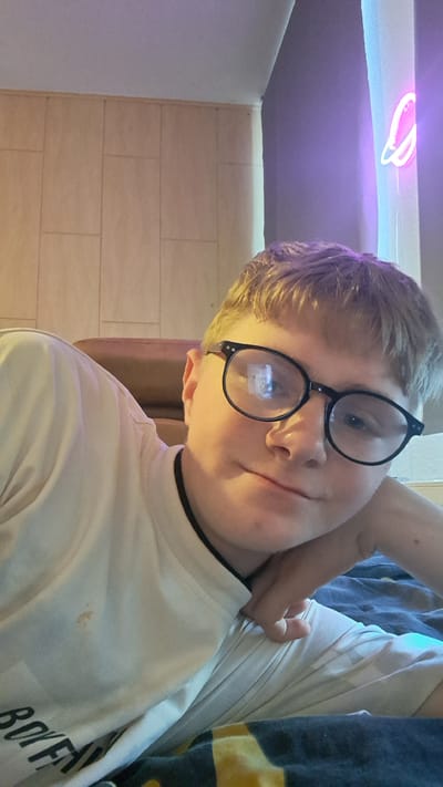 This media file is a close-up, low-angle shot of a young person wearing glasses and a white t-shirt. The person is leaning their head on their hand, looking downwards and slightly to the left. Their hair is blonde and styled in short layers. The t-shirt has a black collar and some dark text on the upper left side, which appears to read "BOYF". A small brown stain is visible on the t-shirt, just below the collar.

The background consists of a light-colored, tiled wall. To the right of the person, there is a surface with a purple and blue neon light, shaped like a ring or halo, casting a glow. The overall setting appears to be indoors, possibly a bedroom, given the relaxed posture of the subject and the ambient lighting. The lighting is dim, with the neon light providing a prominent colorful accent. There are no clear indicators of the time of day or weather conditions. The subject's expression is difficult to discern due to their pose and the lighting.