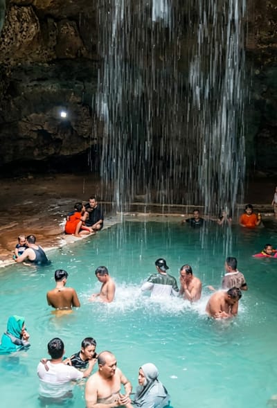 A group of people are bathing in a pool of clear turquoise water in a cave, with a waterfall cascading from above. The cave walls are rocky and textured, with some vegetation visible in the upper left corner. A small, square light fixture is mounted on the cave wall. Several individuals are submerged in the water, some splashing. Others are seated on the ledge surrounding the pool. In the foreground, a man and woman are conversing in the water, while other groups of people are scattered throughout the pool. The overall scene depicts a recreational activity in a natural, enclosed environment.