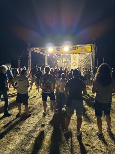 The image shows an outdoor concert at night, possibly in the city of Tutrakan, Bulgaria, given the context. A crowd of people are standing on dry, grassy ground, facing a brightly lit stage. The stage has a large banner behind the performers, featuring a logo and graphics. Several band members are visible on stage, playing instruments and singing. The lighting on stage is a mix of bright spotlights and colored lights, casting beams into the night sky. Lens flares are prominent in the upper portion of the image due to the stage lights. The crowd appears engaged, with some individuals holding up phones. The overall atmosphere suggests a lively musical event.