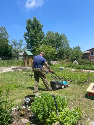 A man wearing a blue t-shirt and camouflage pants is mowing a grassy lawn with a light blue and black lawnmower. He is wearing a camouflage baseball cap. The lawnmower has the brand name "Oasis" visible on it. He is standing in a yard with trees and bushes in the background. There is a small wooden shed with a green roof behind him. A brick house with a balcony is visible to the right of the image. The sky is clear and blue. The scene appears to be a residential area.