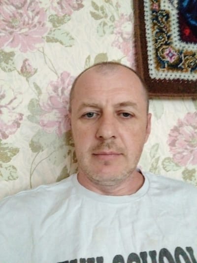 The image is a close-up, selfie-style photograph of a bald, middle-aged man with light skin and stubble. He is wearing a light-colored t-shirt. The man's head and shoulders are visible, and he is looking directly at the camera. The background consists of floral wallpaper with large pink and green flowers. To the right of the man, a portion of a framed tapestry or rug with geometric patterns and colors of blue, red, and brown is visible. The lighting appears to be from an overhead source, casting some shadows on the man's face and shirt. The overall setting suggests an indoor environment, likely a home in Алекшыцкі сельскі Савет, Belarus.