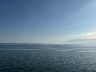 The image displays a vast expanse of dark blue ocean meeting a pale blue sky. The horizon line is sharp and clear, dividing the scene horizontally. Gentle waves create subtle patterns across the water's surface. The sky is mostly clear, with a few wispy clouds scattered sparsely, particularly towards the right side of the frame where a larger, lighter cloud formation is visible. The overall impression is one of calm and open space. The lighting suggests it is daytime, likely late morning or early afternoon, given the brightness and clear sky. There are no discernible people or objects in the frame, making the focus solely on the natural elements of the sea and sky. The location is unknown, but the vastness of the ocean suggests an open coastal or offshore environment. The mood is serene and tranquil. No text is visible in the image.