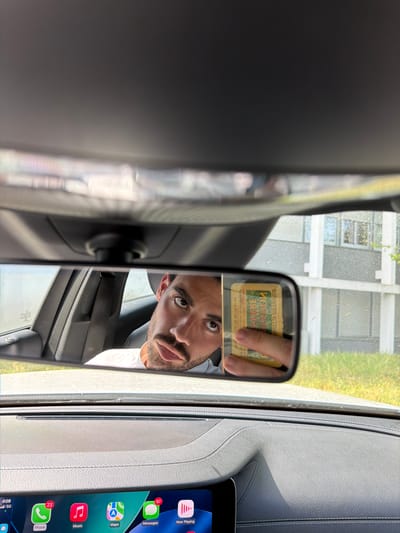 The image shows the interior of a car, specifically the rearview mirror reflecting a man holding a parking validation sticker. The car's dashboard and a digital tablet displaying app icons are visible on the right side of the image. Through the car's windows and in the rearview mirror's reflection, a building and greenery are seen. The parking sticker, reflected in the mirror, mentions "Mont-de-Marsan". The man is holding the sticker in front of the mirror.