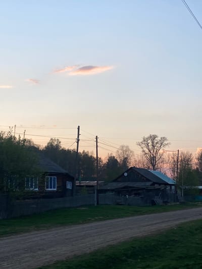 The image presents a rural scene in Узяр, Russia, at dusk. Two wooden houses are visible, weathered and dark, with window frames painted blue. An unpaved road runs alongside the houses, bordered by green grass and foliage. Utility poles with power lines stretch across the frame, silhouetted against the sky. The sky itself is a gradient of light blue to orange, with wisps of cloud. A satellite dish is mounted on one of the houses. Trees with bare branches are interspersed with the houses.