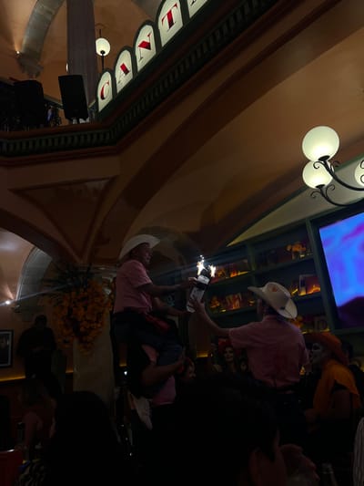 The image depicts an indoor nighttime or dimly lit event within a multi-level establishment, identifiable by a partial illuminated sign reading "CANTA," consistent with a cantina or venue in Guanajuato City, Mexico. The architecture features arched ceilings, large pillars, and a mezzanine level with dark railings, where two large dark speakers are visible. Illumination is provided by wall-mounted globular lamps and recessed lighting.

In the foreground, a performance involves two adult males. One male, wearing a light pink button-up shirt and a white cowboy hat, stands on the shoulders of a second male, also in a similar shirt. The top male holds a dark bottle from which two sparkler-like devices emit bright light and sparks. He is extending this bottle towards the second male below him.

To the left, a prominent floral arrangement composed of orange flowers is visible next to a dark-clothed individual. To the right, a dark shelving unit displays bottles and small decorative items, above which a large screen displays abstract blue and purple imagery. Multiple patrons are present, some of whom are in festive attire or costumes, including at least one individual with skull-like face paint and another with a large orange headdress, suggesting a themed celebration. The lower portion of the image shows indistinct dark forms of the audience.