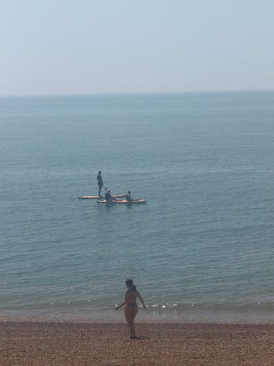 The image shows a shoreline scene. Three individuals are on a wooden structure in the water, seemingly connected by crossbeams. A fourth person is walking in the shallow water at the edge of the beach. The location appears to be a beach in the Folkestone and Hythe District, United Kingdom, based on the beach composition and the sea color.