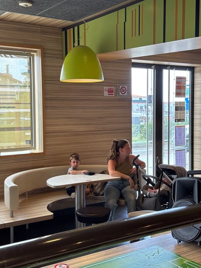 This media file captures an indoor scene, likely a family-friendly restaurant or play area, with a distinctly casual and relaxed atmosphere. The primary subjects are two individuals and a child. A woman, wearing jeans and a tan top, is seated and looking towards a child who is leaning over a white, curved surface. The child, a boy with light brown hair, appears to be interacting with something or someone out of frame. Behind them, a stroller is partially visible, suggesting a family outing.
The setting features wooden paneling on the walls and a large window that offers a glimpse of the outside, hinting at daylight. Reflections on the window suggest an urban or suburban environment with buildings and vegetation. A prominent bright green, bell-shaped light fixture hangs from the ceiling, adding a splash of color to the scene. The overall aesthetic is modern and clean, with smooth surfaces and a functional design.
Two circular signs are visible on the wall. One contains a red circle with a diagonal line through it and a symbol that could indicate a prohibition, possibly related to smoking. The other sign is red and white and features a logo that is difficult to decipher but appears to be a brand or establishment marker, possibly a McDonald's logo given the common association of such play structures with the fast-food chain.
The lighting in the image is bright, suggesting it was taken during the day. The mood is one of everyday life, with no strong indicators of specific emotions beyond casual engagement. The perspective is slightly tilted, adding a dynamic feel to the composition. The presence of the stroller and child indicates a family environment where people are likely dining or taking a break.
