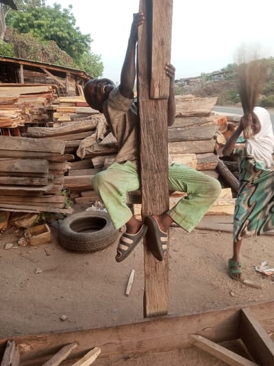 A boy in green pants, a striped shirt, and sandals hangs from a wooden post, smiling and looking up. His arms are extended above his head, gripping the post. A girl in a white headscarf and patterned skirt stands nearby, holding a bundle of sticks resembling a broom. Wood piles, a tire, and various containers surround them. The setting appears to be a dusty outdoor area with some buildings visible in the background.
