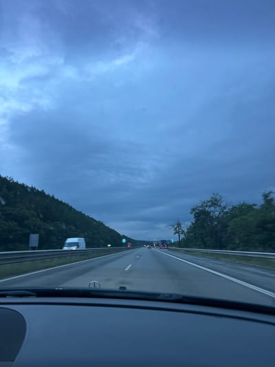 The image depicts a view from inside a vehicle, looking out onto a highway in Novi Han, Bulgaria. The highway stretches into the distance, with multiple lanes visible. Trees and a hillside covered in lush green foliage line the left side of the road, while the right side appears to be the shoulder and interior of the vehicle.

Several vehicles are on the highway, including trucks and cars, suggesting ongoing traffic. Traffic signs, including one with a blue circular background and a white arrow pointing left, are visible along the roadside. The sky is overcast and a deep blue, indicating either early morning or late evening, or possibly impending rain. The overall lighting is dim, consistent with twilight.

No people are clearly visible. The primary focus is the highway scene and the natural landscape. The mood conveyed is one of travel, perhaps a journey through a rural or semi-rural area as dusk approaches. There is no visible text that can be read with certainty.