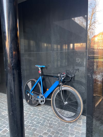 An urban outdoor scene in Uccle-Ukkel, Belgium, depicts a single bicycle leaning against a dark, possibly granite or tiled, building facade. The bicycle features a bright blue main frame triangle and a black top tube, seat post, and front fork. It is equipped with drop handlebars, a light blue saddle, a silver crankset, and a single-speed or fixed-gear drivetrain. The rear wheel is a black spoked disc wheel, while the front wheel is a black, deep-section carbon wheel with tan-colored sidewall tires.
To the immediate left of the bicycle, a large, dark cylindrical pillar extends vertically, partially obscuring the view of the wall. The ground beneath the bicycle and pillar consists of grey, irregularly shaped cobblestone paving.
The dark facade exhibits reflective properties, showing distorted reflections of an outdoor environment. These reflections include bare trees, the warm-toned light of what appears to be a low sun, and distant buildings with reddish-orange rooftops. A vertical glass or highly reflective panel is visible on the far right, further reflecting the outdoor scene. No individuals are present in the image.