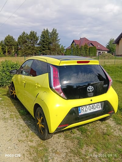 A yellow Toyota Aygo is parked on a gravel and grass area in Wilczyce, Poland. The car's rear is facing the camera, showing its license plate RZ 0404S and a "TOYOTA" emblem on the black rear panel. The car has a black roof and black accents around the rear window and bumper. The surrounding environment includes green bushes to the left, a grassy field with a fence in the background, and trees and houses further in the distance under a cloudy sky. The timestamp "09.06.2026 09:15" is visible in the lower right corner, along with "POCO C75" in the lower left.