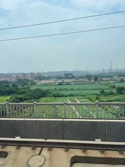 The image depicts a landscape scene in Dongguan, China, viewed from an elevated perspective. The foreground features a metal railing, suggesting the viewpoint is from a balcony, bridge, or similar structure. Beyond the railing, a vast expanse of agricultural fields, likely rice paddies or lotus fields given the characteristic greenery and water channels, stretches into the distance. Interspersed within the fields are trees and small structures.
Further into the background, a cityscape emerges with numerous multi-story buildings, indicating an urban or semi-urban area. Dominating the skyline are several large electricity pylons, a common sight in many developed regions. The sky is overcast with light clouds, suggesting it is daytime, possibly mid-morning or afternoon, with diffused light. The overall atmosphere is somewhat hazy, which is not uncommon in industrial or densely populated areas.
No people are clearly visible in the frame, and the focus is on the natural and built environment. The scene conveys a sense of expanse and the coexistence of agricultural land with urban development. The details of the railing suggest a functional, possibly modern, structure. A small white rectangular label with faint text is visible on the dark paneling behind the railing, but the text is illegible.