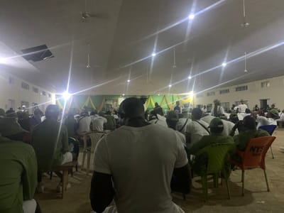 The image displays a densely populated indoor hall in Kubi A, Nigeria, where approximately 60-80 individuals, predominantly young adults, are seated and facing forward. The majority of attendees are dressed in olive green and white attire, consistent with National Youth Service Corps (NYSC) uniforms, including olive green caps, white t-shirts, and olive green trousers or shirts. Seating arrangements consist of various plastic chairs, one orange chair explicitly labeled "NYSC HALL," and several wooden benches.
The hall features light-colored walls and a high ceiling equipped with multiple fluorescent light fixtures and several white ceiling fans. A section of the ceiling exhibits a missing or damaged panel, exposing a dark void. The focal point at the front of the hall is adorned with green and yellow fabric drapes and is intensely illuminated by several bright artificial lights, creating prominent lens flares. A large screen or banner is visible to the right of this decorated area. The floor is a light-colored, possibly concrete or tiled surface. The assembled group suggests participation in a formal gathering, lecture, or event.