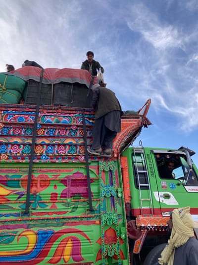 The image depicts a highly decorated cargo truck in Chaman City Tehsil, Pakistan, under a sky with scattered cirrus clouds. The truck's body is predominantly bright green, adorned with intricate, multi-colored floral and geometric "truck art" patterns in red, orange, blue, yellow, pink, and purple. The truck is heavily laden with cargo, much of which is covered by a red tarpaulin and secured by dark straps or ropes. Green bundled items are also visible on the left side of the cargo bed.
Four individuals are present. One man, wearing a light shirt and dark pants, stands atop the cargo, holding a white object in his right hand, looking towards the viewer. Below him, another man, dressed in dark traditional attire and a light cap, is actively climbing the side of the truck's cargo bed using a ladder-like structure. A third man is visible smiling through the open driver-side window of the truck's cab. The cab is green and features text including "HINO," "Ranger," "EURO," and Chinese characters. A metal ladder is attached to the side of the cab. A fourth individual, partially visible in the lower right foreground, wears a light-colored head covering and dark clothing, positioned near the front of the truck. The scene suggests activities related to loading, unloading, or preparing the truck for transit.