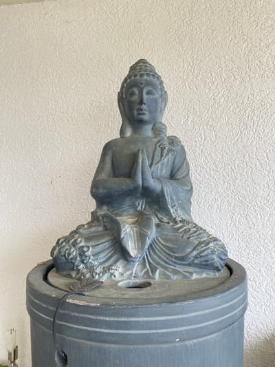 A stone Buddha statue is affixed to a round, grey, horizontal object with ridges, possibly a decorative fountain element, set against a textured white wall. The image appears to be taken indoors. There are no people visible. There is no location in the city of Trebesin, Montenegro.