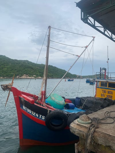 The image depicts a fishing boat moored at a pier in Vĩnh Hải, Vietnam. The boat, painted red and blue with black accents, has the registration number "NT90124 TS" visible on its side. Various objects are present on the boat, including blue and green buoys, a red crate, and spare tires used as fenders. Ropes and rigging are prominent, indicating its operational status.
In the background, several other boats are visible in the water, suggesting a fishing harbor. A lush, green, hilly island rises from the water behind the boats, with scattered rocks and vegetation. The sky is overcast with clouds, hinting at potentially cloudy or impending weather. The overall scene suggests a quiet moment, likely in the morning or afternoon, as the boats are docked and the day's fishing activities may be paused or concluded. The location context of Vĩnh Hải, Vietnam, is supported by the distinctive green, rocky island topography common in coastal areas of Vietnam. There is no visible activity of people.