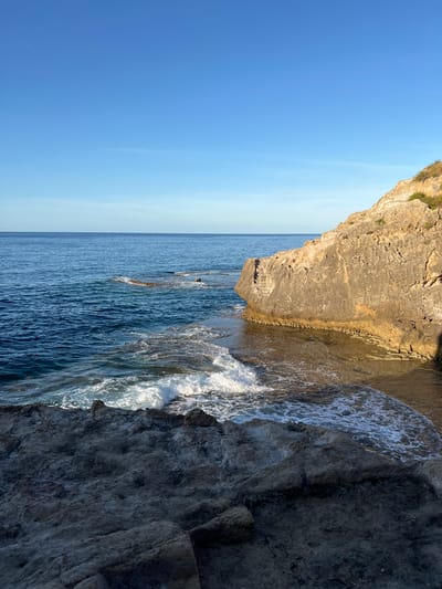 The image depicts a rocky coastal scene in Sperone, Italy, under a clear, transitioning blue sky. The upper portion of the frame features a cloudless sky, light blue at the horizon and deepening overhead. Below, the calm, deep blue sea extends to the horizon, with gentle waves breaking on the foreground and midground rocks. In the foreground, dark, textured, irregular rock formations occupy the lower left and central areas. To the right, a prominent, light brown, stratified cliff face rises, adorned with sparse green vegetation on its upper edge. The base of this cliff meets the water, forming a shallow, light-colored inlet where gentle ripples are visible. No individuals, animals, or man-made objects are present in the scene.