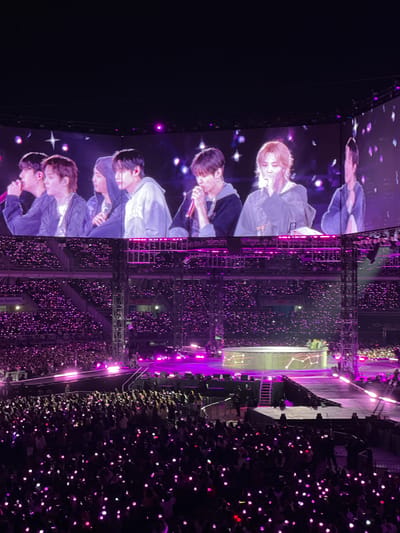 Here's a description of the image:

The image captures a concert scene, possibly in Goyang-si, South Korea given the prompt. A large screen on the left displays five people. The audience is visible in the center, holding up lights which fill the arena with a purple hue. The stage extends to the right, with additional people visible. A smaller screen is placed behind the stage, with the display appearing in white and black colors. The scene appears to be from a large concert.