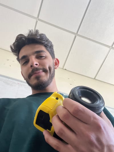 A low-angle, close-up shot captures a young man from the chest up, holding photography equipment. He has dark, wavy hair, a dark beard and mustache, and prominent dimples. The man is wearing a dark green long-sleeved shirt. He is holding a black camera lens in his right hand and a yellow object with a ribbed texture and a black grip in his left hand. The background consists of a white acoustic tile ceiling with a grid pattern of white beams. The overall lighting is bright and even.