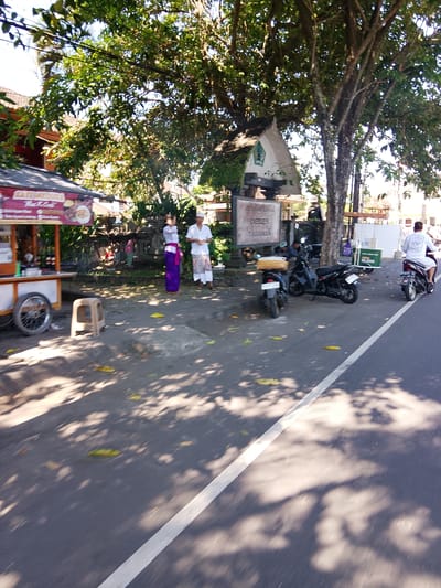 The image depicts a street scene in Tabanan, Indonesia, under a bright, sunny sky with dappled shade from overhead trees. In the foreground, a paved road with a white line marking the lane is visible, with shadows of leaves and branches cast across it. To the left, a food cart with a red and white awning is parked, displaying a sign with Indonesian text and images of food. Beside the cart, a small, light-colored stool sits on the ground.

Further back, two individuals are standing near a traditional Balinese stone wall. One person is wearing a long purple sarong and a white top, while the other is in a white shirt and patterned sarong. Behind them, a small, white structure with a green roof and a triangular entrance is partially visible.

Several motorcycles are parked along the side of the road. Two are black, one in front of the other, with one having a yellow container on its rear. Another black motorcycle is parked further down, with a rider wearing a white shirt and pants, facing away from the camera. A white cooler box with the number "10.000" on it is placed near the motorcycles. A signpost with Indonesian text, including "DEBES" and "TABANAN," is also visible. The background shows more trees, foliage, and glimpses of buildings, suggesting a roadside setting within the city of Tabanan.