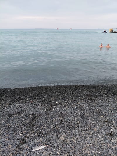 A wide shot shows a pebble beach in Ақши, Kazakhstan, with the calm, gray water of a large body of water in the background. Two people are partially submerged in the water, with their upper bodies visible. In the distance, a jet ski is being ridden by a person. Further out, two small boats and a larger vessel are present. A wooden pier extends from the right side of the frame into the water. The sky is overcast. A single white feather lies on the pebble beach in the foreground.