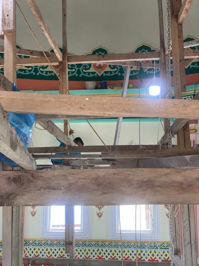 The image depicts the interior of a building, likely a mosque or community hall, undergoing renovation or construction in Mns Asan, Indonesia. The perspective is from below, looking upwards through a dense, multi-layered wooden scaffolding structure that occupies much of the foreground and midground.
One individual is partially visible in the upper-mid section of the image, positioned behind a wooden beam. This person appears to be male, wearing a blue shirt, with only their head and upper torso visible, engaged in an action involving their hands near what appears to be a wire or rope.
The interior walls and ceiling are painted in light green and mint hues. Along the upper wall, near the ceiling line, a decorative band features a green and white scrolling pattern with pink floral accents, bordered by a peach-colored stripe below and a light green stripe above. Further down the wall, below the main scaffolding, a second, more vibrant decorative band is visible, composed of a repeating geometric pattern of red, yellow, blue, green, and white shapes.
Two rectangular windows, emitting bright natural light, are visible in the lower background, set within white or light-colored frames, below the lower decorative band.
On an elevated ledge in the upper background, several objects are present: two white bowls or containers, a blue patterned container, and some yellow fabric or rags. A single, bright white LED light bulb hangs from a wire near the right side of this elevated area. Blue tarpaulin is wrapped around a wooden beam on the left side of the scaffolding, and metal chains hang from beams on the right. Yellow ropes or wires are also visible crisscrossing the scene.