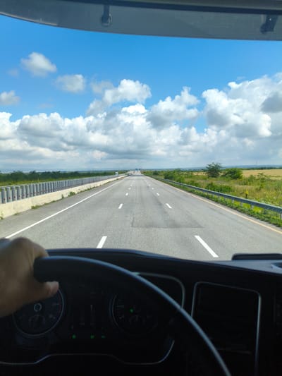 The image shows a driver's perspective from inside a vehicle, looking out at a highway. The driver's left hand is visible on the steering wheel. The road stretches ahead, with white lane markings. On the left side of the road, there is a concrete barrier with vertical gray posts. On the right side, there is a metal guardrail and vegetation. The sky is blue with scattered white clouds. The scene appears to be a highway in a rural or semi-rural area. There are no discernible landmarks that would place the image specifically in Beslan, Russia.