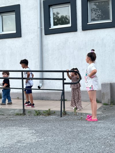 Four children are standing in front of a white building with black-trimmed windows. On the left, a young boy in blue jeans and a black t-shirt with orange trim is holding a white object. Next to him, a slightly older boy in a white jersey with navy shorts and black and red sneakers stands with his hands on his hips. Behind them, a young girl in a leopard-print dress is holding onto a black railing. To the far right, a girl in a white t-shirt with a pink graphic and pink shorts stands with her hands on her hips, wearing bright pink slides. The building appears to be a commercial or residential property, with a drainpipe running vertically between the windows. The ground in front of the building is paved with light-colored tiles, and a gravel area is in the foreground. The overall scene suggests an outdoor gathering or a moment of observation.
