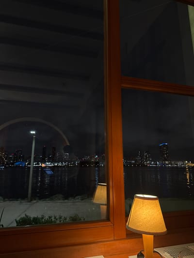 A wood-framed window reveals a nighttime cityscape across water. Lights from buildings and reflections shimmer. A lamp with a tan shade sits on the right edge of the frame, emitting a warm glow. To the right of the lit lamp is an air conditioner and wood framing. The window frame is thick and dark wood.
