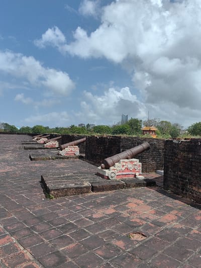 The image displays an outdoor historical fortification, specifically a cannon battery, situated in Phú Xuân Ward, Vietnam, under a bright blue sky partially covered by scattered white cumulus clouds. Five dark, weathered cannons, likely constructed of cast iron, are arranged in a linear sequence, extending from the foreground towards the midground. Each cannon rests on a light-colored, ornate base featuring red trim and carved patterns. These cannons are positioned behind a series of low, dark reddish-brown brick walls, forming individual defensive embrasures. The ground is paved with square, weathered reddish-brown tiles or bricks, showing minor discoloration and a small circular depression in the immediate foreground. A dense line of green trees spans the horizon in the background. Above the tree line, a modern cityscape is visible, characterized by multiple buildings, including a tall, multi-story structure with an angular roofline on the right side. To the left of this skyscraper, a smaller, traditional Vietnamese pavilion with an orange roof and yellow walls is also visible. No individuals are present in the scene.