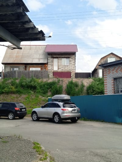 This image captures an outdoor scene on a cloudy day, possibly in Pavlovsk, Russia, with a focus on residential buildings and vehicles. In the foreground, a grey asphalt surface transitions to a gravel and grassy area. Two SUVs are parked on the asphalt. A black SUV is on the left, and a silver SUV is to its right, equipped with a roof-top cargo carrier.

Behind the vehicles, a raised grassy embankment is covered in dense green foliage. A weathered wooden fence runs along the top of the embankment. Beyond the fence, two houses are visible. The house on the left features a corrugated grey roof and wooden plank siding with a white-framed window. Adjacent to it, a smaller house is constructed of cinder blocks with a red-tiled roof and a white-framed door and window. Further to the right, another house, partially obscured, is built with red and grey bricks and has a sloped wooden roof with a window. A tall, blue corrugated metal fence runs horizontally across the midground, separating the embankment from the houses.

Overhead, power lines are strung across the sky, and a light fixture is mounted on the underside of a wooden overhang in the far left of the image. The sky is partly cloudy, with patches of blue visible.
