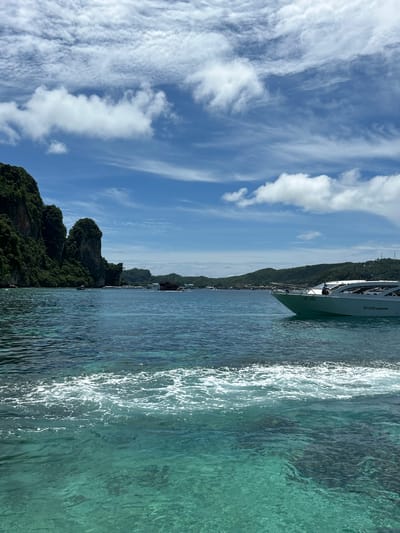 The image depicts a scenic view of Haad Hin Khom, Thailand, captured from the water. The scene is dominated by a bright blue sky filled with wispy white clouds, suggesting a clear and sunny day, likely during daylight hours. To the right of the frame, the turquoise water is crystal clear, with sunlight creating shimmering reflections on its surface. The water appears calm, with gentle ripples.
In the mid-ground, a lush, green mountainous landscape rises from the water's edge. The slopes are densely covered with vegetation, typical of a tropical island environment. Scattered along the coastline are small buildings and boats, indicating a populated area and a hub for maritime activity.
A white boat is prominently featured in the upper right portion of the image, positioned close to the viewer and appearing to be in motion, as evidenced by the wake trailing behind it. The boat has a sleek, modern design. Visible text on the boat reads "LOVE andaman" and a smaller line of text below it.
The overall atmosphere is serene and idyllic, evoking a sense of vacation and natural beauty. The combination of the vibrant blue sky, clear water, and verdant landscape points to a desirable travel destination.