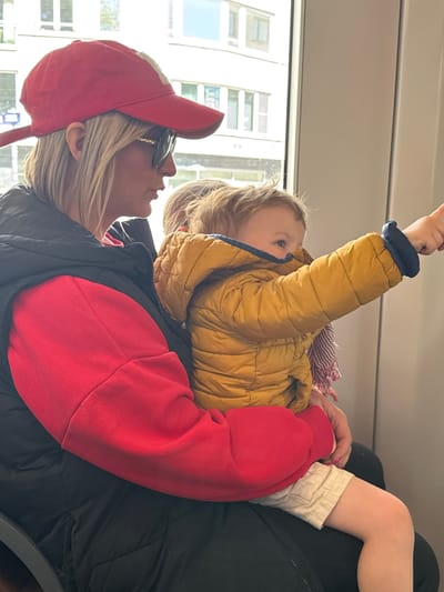 The image captures a candid moment between an adult and a young child inside what appears to be public transportation, likely a train or tram, given the large windows and interior structure. The scene is set during the daytime, with bright light illuminating the urban buildings visible through the windows.

The adult, possibly a woman, is seen from the side, wearing a red baseball cap, dark sunglasses perched on her head, a bright red long-sleeved top, and a dark puffer vest or jacket. She has blonde hair and a faint smile, gazing upwards and slightly to her left. Her fingernails are painted light pink.

A young child, appearing to be a toddler or preschooler, is nestled on the adult's lap or chest. The child is dressed in a yellow puffer jacket and has a striped scarf visible around their neck. The child's blonde hair is also visible. They are looking upwards and to their right, with their right arm raised and hand extended, seemingly pointing or reaching for something overhead. Part of the child's bent left leg, wearing light-colored shorts or pants, is also visible.

The overall impression is one of gentle interaction or shared observation, perhaps the child is pointing out something interesting, and the adult is engaged. There is no visible text in the image.