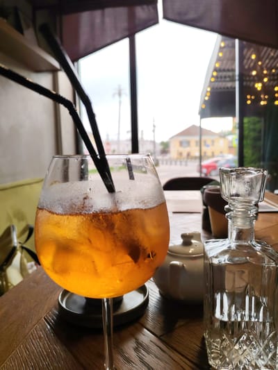 A close-up, eye-level, indoor shot showcases a glass of amber-colored liquid with ice and two black straws, positioned on a dark wooden table. To the right of the glass, a clear glass decanter with a decorative base and stopper is visible, along with a small white teapot. The background, viewed through a large window, reveals an outdoor scene with buildings and a street, suggesting a restaurant or cafe setting in the city of Selivanovo, Russia. The lighting is soft and diffused, with bokeh effects from string lights visible in the upper right portion of the background.