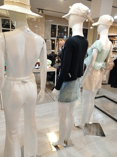 The image shows three mannequins in a clothing store located at Latitude: -6.763, Longitude: 111.03 in Kecamatan Margorejo, Indonesia. The mannequins are dressed in different outfits and are standing on square platforms. The first mannequin on the left is wearing a white outfit with a wide brimmed white hat. The second mannequin is wearing a black sweater, a denim skirt, and a white hat. The third mannequin is wearing a light blue and pink outfit and a white hat. The mannequins are standing on tiled floors. The store appears to be well-lit with multiple lights. A sign is visible on the right side of the image with a price tag. There are additional mannequins and clothing racks in the background.