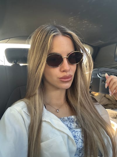 A young woman is sitting in the driver's seat of a car, her head tilted slightly to the left. She is wearing large, oval-shaped sunglasses with gold frames and a light-colored collared shirt over a blue and white patterned top. A delicate silver necklace with a small pendant rests on her collarbone. Her blonde hair, with subtle highlights, falls past her shoulders, framing her face. Her lips are slightly parted, and her left hand is visible, resting near her face, showcasing her manicured nails adorned with small, clear jewels. The interior of the car is dimly lit, with sunlight dappling through the windows onto the ceiling. The background is out of focus, showing the dark interior of the car and a glimpse of the seat behind her. The car appears to be stationary, possibly parked, with the sunlight suggesting it is daytime. The overall impression is a candid, relaxed moment captured within the vehicle, potentially in or around the city of Strumyani, Bulgaria, given the contextual information provided.