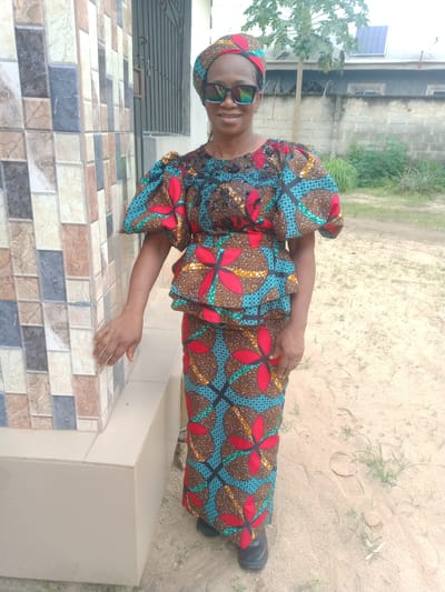 A woman is featured prominently in a full-length outdoor portrait, standing and smiling slightly. She is dressed in a vibrant, matching traditional African print ensemble, which includes a headscarf, a peplum-style top with distinct puffed sleeves, and a long skirt. The fabric features intricate patterns in shades of brown, red, teal, and black. She wears dark sunglasses and black, closed-toe shoes. Her left hand is gently placed on a decorative tiled wall composed of multi-colored square tiles in brown, grey, blue, and cream hues, resting on a light-colored ledge.
The setting appears to be a yard or outdoor area beside a building in Obehie, Nigeria. In the immediate background to her left, a window with metal bars is visible. To her right and in the farther background, the ground is sandy with sparse patches of green grass and weeds. A small tree, possibly a papaya tree, with green fruit, stands behind her. Further back, there is a low cinder block wall, and beyond that, another building with a corrugated metal roof on which a solar panel is mounted. The scene is brightly lit, indicating it is daytime, under clear or sunny weather conditions. No discernible text is visible in the image.