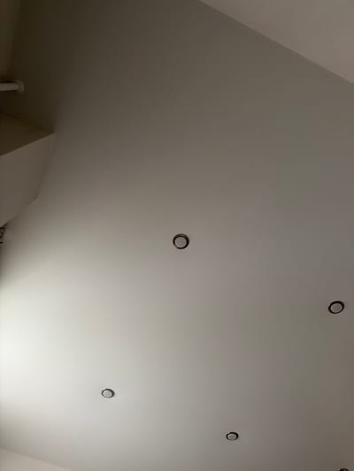 The image shows a white ceiling with several recessed circular light fixtures. One light fixture is visible in the top center, another in the middle right, and two are visible in the bottom left and bottom center. The ceiling appears to be sloped in the upper left corner, where a portion of a wall or architectural feature is also visible. There are no people or visible actions in the image. The lighting is dim, with shadows cast by the light fixtures and the architectural elements. The scene is devoid of any context related to the city of Crokhovo, Russia.
