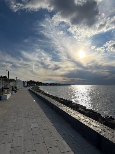 This image, set in Taman, Russia, captures a modern coastal promenade under a dynamic, partly cloudy sky. The sun, bright and prominent, is high in the sky, indicating midday or early afternoon, and its light creates a dazzling, shimmering effect on the surface of the large body of water (likely a sea or large lake) on the right. This water meets a dark, rocky shoreline, which is neatly bordered by a dark grey concrete wall or barrier.

On the left, a wide promenade, paved with uniform grey rectangular blocks, stretches into the distance. Along this walkway, several dark, modern streetlights or poles are visible, as well as white, minimalist benches. A few people are scattered along the promenade, some walking leisurely, while others are seated on the benches, appearing relaxed and enjoying the pleasant outdoor setting. Further along, a contemporary fence or glass barrier separates the promenade from a more verdant area with distant buildings, hinting at a nearby town or settlement.

The sky is a notable feature, displaying a mix of large, fluffy white clouds, darker grey formations, and delicate wisps of cirrus clouds against a clear blue expanse, suggesting fine weather. The scene conveys a sense of tranquility and an inviting atmosphere for outdoor activities. No visible text is present in the image.