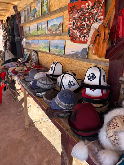 Captured at an angle, the scene depicts a vendor's booth in Tosor, Kyrgyzstan. The booth's roof is constructed of plywood and wood supports, showcasing an array of items for sale. Hats, some with intricate embroidery and fur trim, dominate the view. Leather bags of various styles and colors, as well as small landscape paintings, are also displayed. Slippers and other miscellaneous items are arranged along the lower edge of the booth. The ground below the booth is dirt.