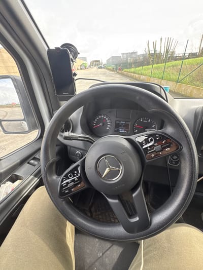 The image depicts a first-person view from the driver's seat of a Mercedes-Benz commercial vehicle, likely a van, situated on an asphalt industrial road in Roeselare, Belgium. The black multi-function steering wheel, bearing the Mercedes-Benz logo and an "AIRBAG" inscription, occupies the central foreground. The dashboard instrument cluster shows the vehicle is stationary, with the speedometer at 0 and the tachometer at idle, while a central digital display reads "13:01," "25131" (trip mileage), and "186952" (total mileage). A Honeywell-branded suction cup mount is affixed to the windshield, holding a dark-colored smartphone vertically in the upper left quadrant. The driver's legs, clad in light brown trousers, are visible in the lower portion of the frame. Through the windshield, an industrial landscape unfolds under an overcast sky. To the left, a large light-colored industrial building is partially visible, with its reflection in the side mirror. To the right, a green metal fence separates the paved area from a grassy strip, beyond which industrial structures and stored materials, including tall cylindrical objects, are present.
