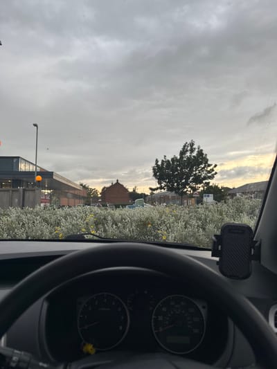The image shows a view from the driver's seat of a car. The car is parked in a parking lot, facing a row of buildings, most of which are obscured by a bush of white flowering plants in the foreground. A single tree can be seen in the distance, behind the buildings, and the sky is overcast with a light dusting of clouds. The car is parked close to the curb and the car dashboard is visible, including the steering wheel, the dashboard, and a black phone holder mounted on the windshield. The speed gauge on the dashboard reads zero. The image was taken in Pocklington, a town in the East Riding of Yorkshire, England, and is likely located on or near York Road. The buildings in the distance are likely part of a commercial or industrial development. The view from the car suggests that it is a late afternoon or early evening.