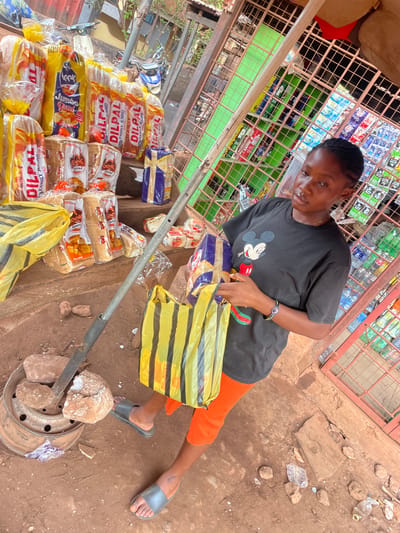 A woman stands near a roadside stall in Otukpa, Nigeria, packaging bread. The stall is a simple metal-framed structure filled with various goods. The woman wears a dark t-shirt with a Mickey Mouse graphic and orange shorts, and she is packing loaves of DILPAL brand bread into a yellow and black striped bag. The ground is dusty and scattered with debris. The stall is stocked with an assortment of items, including bottled drinks and packaged goods. A metal pole rests on the ground, secured by a metal base and rocks.