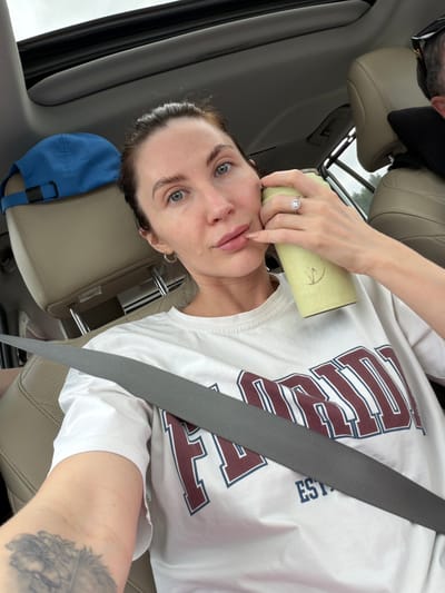 A selfie-style image captures a woman in the passenger seat of a car in Opytnoe Pole, Russia. The photo is taken from a high angle, showing the woman looking directly at the camera with light-colored eyes and dark hair pulled back. She wears a white t-shirt featuring partially visible maroon and blue outlined text that reads "UST" and "EST.". A grey seatbelt crosses her chest. On her left hand, she sports a ring, likely an engagement ring, and has light-colored nail polish. A tattoo is visible on her left arm. She holds a light green cylindrical bottle with a faint symbol near her face. Her expression is neutral with slightly parted lips.

The car's interior has tan/beige leather seats and dark grey trim. A blue baseball cap rests on the headrest beside her. In the upper left, a reflection or partial view of another person, possibly a man wearing sunglasses or a hat, is visible behind a headrest. The daylight visible through the windows suggests an overcast or cloudy sky during the daytime.