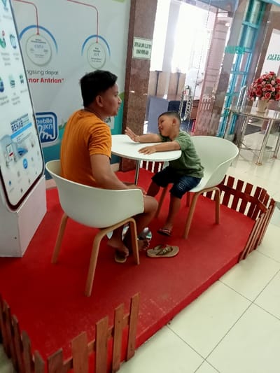 The image shows a man and a young boy sitting at a table in a shopping mall. The man is wearing a yellow shirt and is sitting on a white chair with wooden legs. The boy is wearing a green shirt and blue jeans. He is sitting on a white chair with wooden legs. They are sitting at a round, white table. The table is set on a red carpet surrounded by a small fence made of brown wooden planks. The man is looking at the boy, who is looking at the camera. The man has his feet flat on the red carpet. The boy is wearing sandals and his feet are off the ground, hanging from the chair. There is a large white sign with text in Indonesian and a small sign with a mobile phone. Behind the man and boy, there is a glass wall. In the distance, there is a potted plant and a chair. The location is the Kecamatan Margorejo area of Surabaya, Indonesia, at latitude: -6.76, longitude: 111.015.
