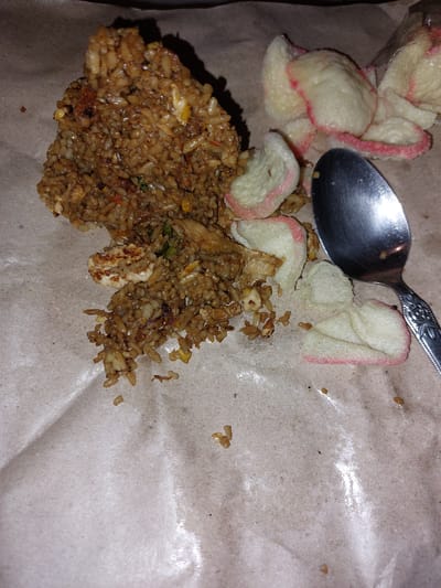 The image shows a close-up, overhead view of a portion of fried rice served on brown paper, likely takeout packaging. A metallic spoon rests on top of the food, its reflective surface catching the light. Scattered around the rice are several light-colored crackers with pink edges, possibly prawn crackers (krupuk). The fried rice appears to be seasoned and contains visible pieces of chicken and other ingredients. The brown paper packaging is slightly creased and shows some oil stains. The lighting is dim, suggesting it might be evening or an indoor setting. There is no visible text in the image. The overall impression is that of a hastily prepared or partially consumed meal, possibly in a casual setting in Karangjati, Indonesia.