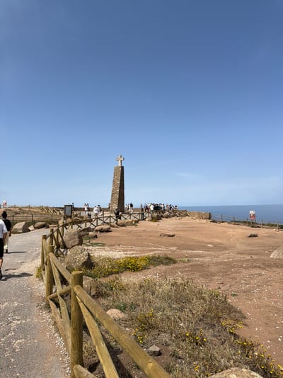 The image depicts Cabo da Roca, the westernmost point of mainland Europe, located near Colares, Portugal. A stone monument with a cross stands prominently in the center, surrounded by a group of tourists. The landscape is characterized by dry, reddish soil, scattered rocks, and sparse vegetation with yellow wildflowers. A wooden fence demarcates a pathway leading towards the monument and a viewing area overlooking the vast blue ocean. The sky is clear and bright blue. Several people are visible walking and observing the scenery, with some gathered around the monument and others near the edge of the cliff. The overall scene suggests a popular tourist destination on a sunny day.