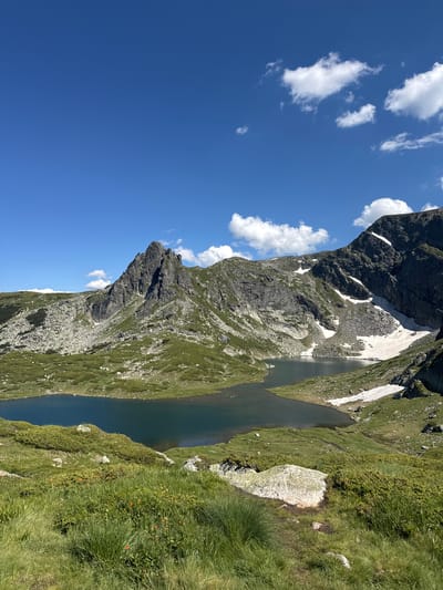 The image depicts a scenic mountain landscape in Panichishte, Bulgaria, featuring a clear blue sky with scattered white clouds. A tranquil, dark blue lake occupies the middle ground, reflecting the surrounding mountains. Steep, rugged mountains rise behind the lake, with patches of snow visible on their slopes, indicating a high altitude. The terrain is covered in lush green grass, interspersed with rocks and low-lying vegetation. In the foreground, a grassy slope leads down towards the lake. The scene is natural and serene, with no visible people or man-made structures. The lighting suggests a bright, sunny day.